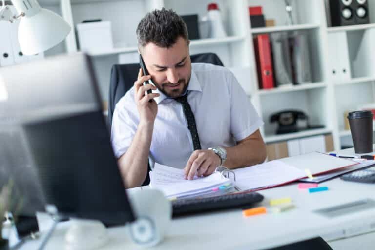 A man is sitting in the office, working with documents and talking on the phone.